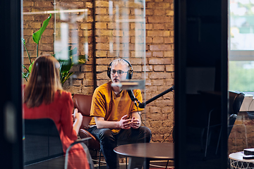 Image showing A gathering of young business professionals, some seated in a glass-walled office, engage in a lively conversation and record an online podcast, embodying modern collaboration and dynamic interaction