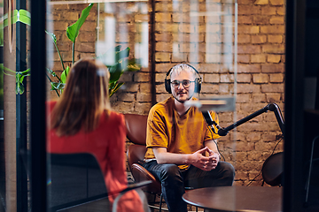 Image showing A gathering of young business professionals, some seated in a glass-walled office, engage in a lively conversation and record an online podcast, embodying modern collaboration and dynamic interaction
