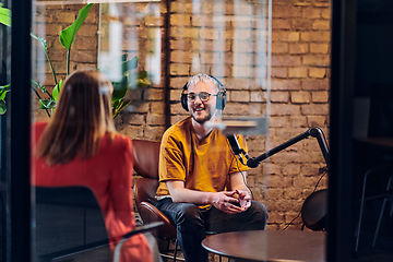 Image showing A gathering of young business professionals, some seated in a glass-walled office, engage in a lively conversation and record an online podcast, embodying modern collaboration and dynamic interaction