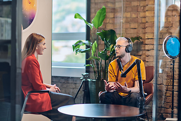 Image showing A gathering of young business professionals, some seated in a glass-walled office, engage in a lively conversation and record an online podcast, embodying modern collaboration and dynamic interaction