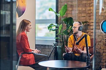 Image showing A gathering of young business professionals, some seated in a glass-walled office, engage in a lively conversation and record an online podcast, embodying modern collaboration and dynamic interaction