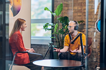 Image showing A gathering of young business professionals, some seated in a glass-walled office, engage in a lively conversation and record an online podcast, embodying modern collaboration and dynamic interaction