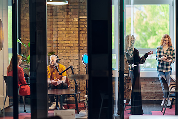 Image showing A gathering of young business professionals, some seated in a glass-walled office, engage in a lively conversation and record an online podcast, embodying modern collaboration and dynamic interaction