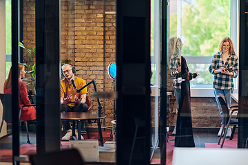 Image showing A gathering of young business professionals, some seated in a glass-walled office, engage in a lively conversation and record an online podcast, embodying modern collaboration and dynamic interaction