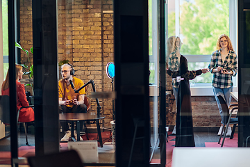 Image showing A gathering of young business professionals, some seated in a glass-walled office, engage in a lively conversation and record an online podcast, embodying modern collaboration and dynamic interaction