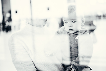 Image showing Thoughtful young mother looking trough window holding his infant baby boy child while waiting to board an airplane at airport terminal departure gates. Travel with baby concept.