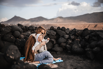 Image showing Mother enjoying winter vacations playing with his infant baby boy son on black sandy volcanic beach of Janubio on Lanzarote island, Spain on windy overcast day. Family travel vacations concept.