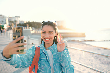 Image showing Smiling, trendy and fun woman, tourist or influencer taking selfie with peace sign gesture on phone for social media, video call or photo. Exploring, traveling and sightseeing at promenade outdoors