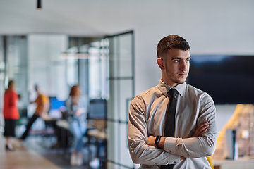 Image showing A young business leader stands with crossed arms in a modern office hallway, radiating confidence and a sense of purpose, embodying a dynamic and inspirational presence.