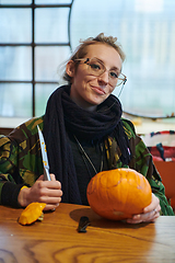 Image showing A modern blonde woman in military uniform is carving spooky pumpkins with a knife for Halloween night