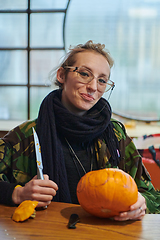 Image showing A modern blonde woman in military uniform is carving spooky pumpkins with a knife for Halloween night