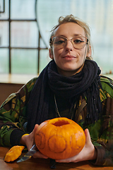 Image showing A modern blonde woman in military uniform is carving spooky pumpkins with a knife for Halloween night