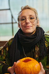 Image showing A modern blonde woman in military uniform is carving spooky pumpkins with a knife for Halloween night