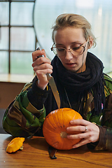 Image showing A modern blonde woman in military uniform is carving spooky pumpkins with a knife for Halloween night