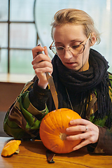 Image showing A modern blonde woman in military uniform is carving spooky pumpkins with a knife for Halloween night