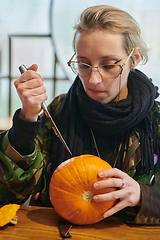 Image showing A modern blonde woman in military uniform is carving spooky pumpkins with a knife for Halloween night