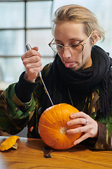 Image showing A modern blonde woman in military uniform is carving spooky pumpkins with a knife for Halloween night