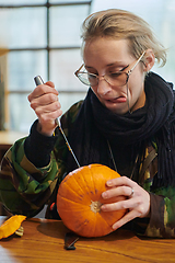 Image showing A modern blonde woman in military uniform is carving spooky pumpkins with a knife for Halloween night