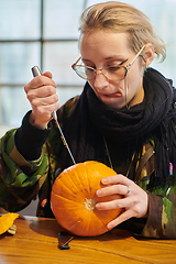 Image showing A modern blonde woman in military uniform is carving spooky pumpkins with a knife for Halloween night