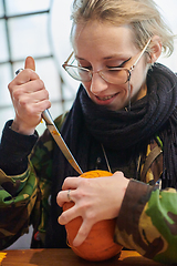 Image showing A modern blonde woman in military uniform is carving spooky pumpkins with a knife for Halloween night