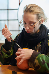 Image showing A modern blonde woman in military uniform is carving spooky pumpkins with a knife for Halloween night