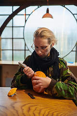 Image showing A modern blonde woman in military uniform is carving spooky pumpkins with a knife for Halloween night