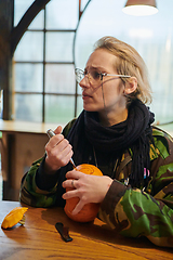 Image showing A modern blonde woman in military uniform is carving spooky pumpkins with a knife for Halloween night