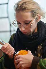 Image showing A modern blonde woman in military uniform is carving spooky pumpkins with a knife for Halloween night