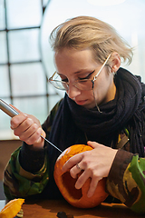 Image showing A modern blonde woman in military uniform is carving spooky pumpkins with a knife for Halloween night