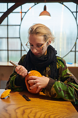 Image showing A modern blonde woman in military uniform is carving spooky pumpkins with a knife for Halloween night