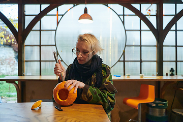 Image showing A modern blonde woman in military uniform is carving spooky pumpkins with a knife for Halloween night