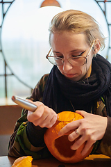 Image showing A modern blonde woman in military uniform is carving spooky pumpkins with a knife for Halloween night