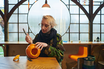 Image showing A modern blonde woman in military uniform is carving spooky pumpkins with a knife for Halloween night