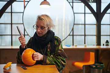 Image showing A modern blonde woman in military uniform is carving spooky pumpkins with a knife for Halloween night