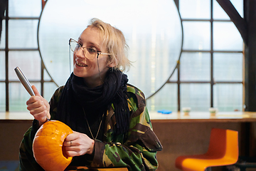 Image showing A modern blonde woman in military uniform is carving spooky pumpkins with a knife for Halloween night