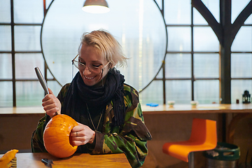 Image showing A modern blonde woman in military uniform is carving spooky pumpkins with a knife for Halloween night