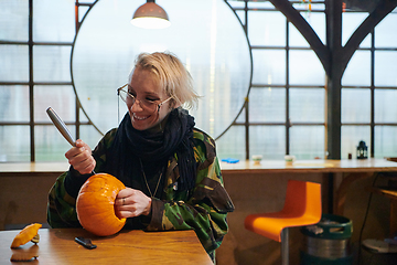 Image showing A modern blonde woman in military uniform is carving spooky pumpkins with a knife for Halloween night