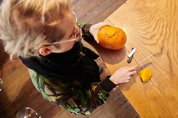 Image showing A modern blonde woman in military uniform is carving spooky pumpkins with a knife for Halloween night