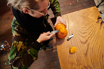 Image showing A modern blonde woman in military uniform is carving spooky pumpkins with a knife for Halloween night