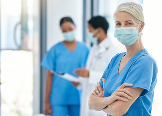 Image showing Success, healthcare and leadership, woman doctor in surgical mask and scrubs, standing arms crossed with coworkers talking in background. Proud, professional and motivated to fight covid at hospital.
