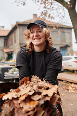 Image showing A stylish, modern young woman takes on the role of a garden caretaker, diligently collecting old, dry leaves and cleaning up the yard in an eco-conscious manner