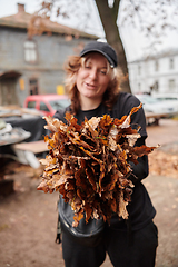 Image showing A stylish, modern young woman takes on the role of a garden caretaker, diligently collecting old, dry leaves and cleaning up the yard in an eco-conscious manner