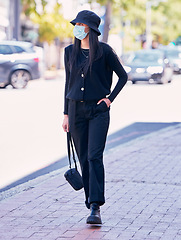 Image showing City woman, on a sidewalk, with a mask on her face and walking down the road. Urban fashion, casual walk and covid protection, life in the new normal. Calm beauty, trendy girl walks on the pavement.