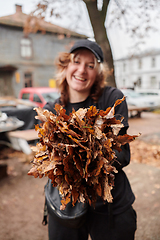 Image showing A stylish, modern young woman takes on the role of a garden caretaker, diligently collecting old, dry leaves and cleaning up the yard in an eco-conscious manner