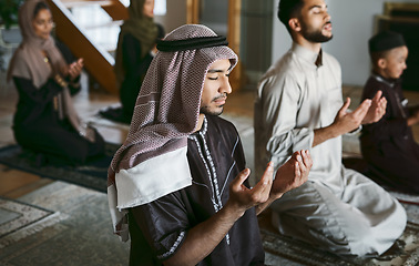 Image showing Young muslim family pray together, sit on knees at home practicing Islam. Religious women wearing hijab and spiritual men wear traditional attire, close eyes and concentrate on prayer with hands up.