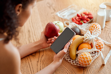 Image showing Colorful, healthy and nutritional organic fruit groceries on table beside woman holding phone and looking online for smoothie recipes. Young lady eating fresh organic apple for wellness vegan diet