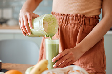 Image showing Woman pouring healthy smoothie in a glass from a blender jar on a counter for detox. Female making fresh green fruit juice in her kitchen with vegetables and consumables for a fit lifestyle.