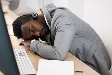 Image showing Tired CRM call center agent sleeping on computer desk at work office with headset on. Exhausted young business man taking a nap on table. African American male feeling burnout resting in workplace.