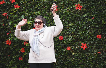 Image showing Funny old woman dancing listening to music on smartphone wearing earphones smiling enjoying fun celebrating retirement in garden with flower wall