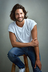 Image showing Style, happy and portrait of man in studio on a stool with casual, cool and stylish outfit. Smile, handsome and confident young male model from Mexico with trendy fashion on chair by gray background.